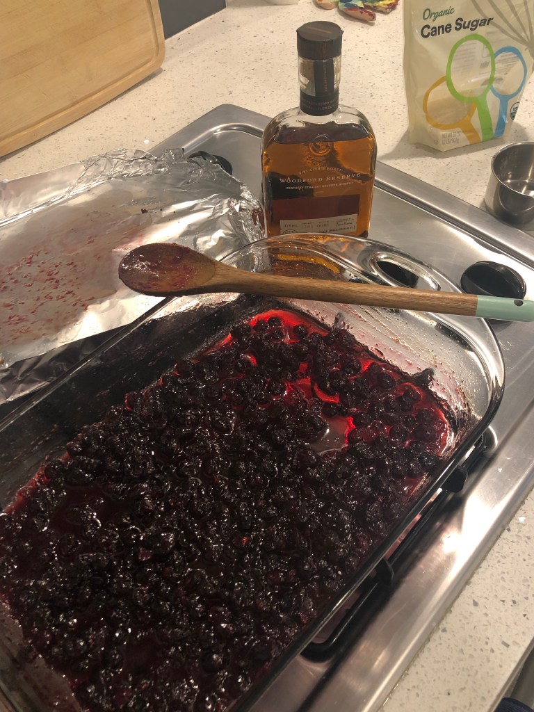 Cranberries in baking dish with wooden spoon lying across one corner and small bottle of bourbon next to it.