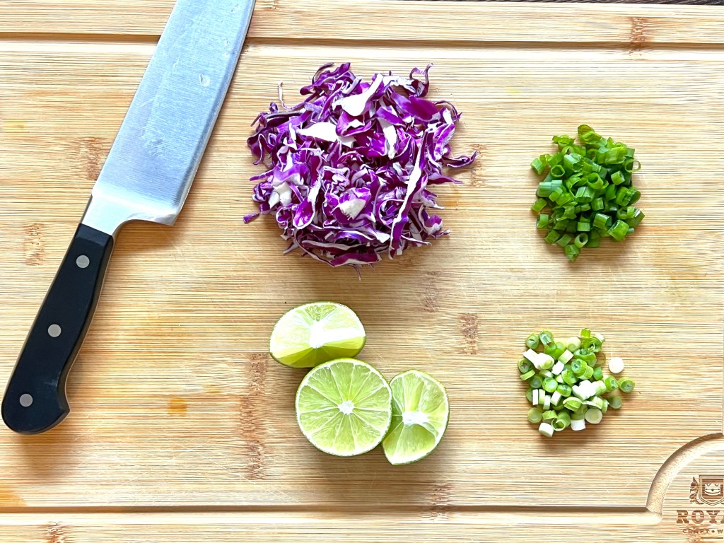 A knife on a cutting board with small piles of sliced purple cabbage. sliced lime, and sliced green onion split into white and dark greens.