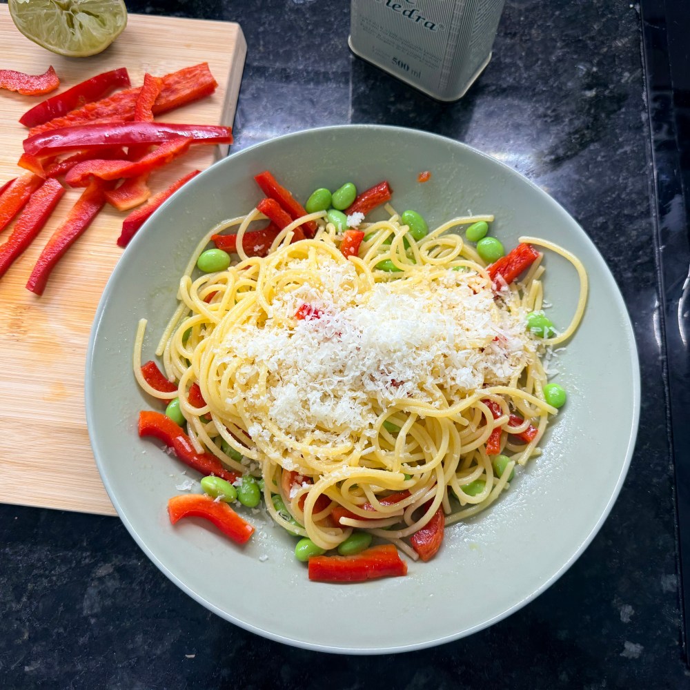 Pale green bowl with spaghetti, edamame, red bell peppers, and covered with grated cheese.